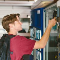 Student teenage boy wearing backpack uses mobile phone to pay for snack and drink at vending machine